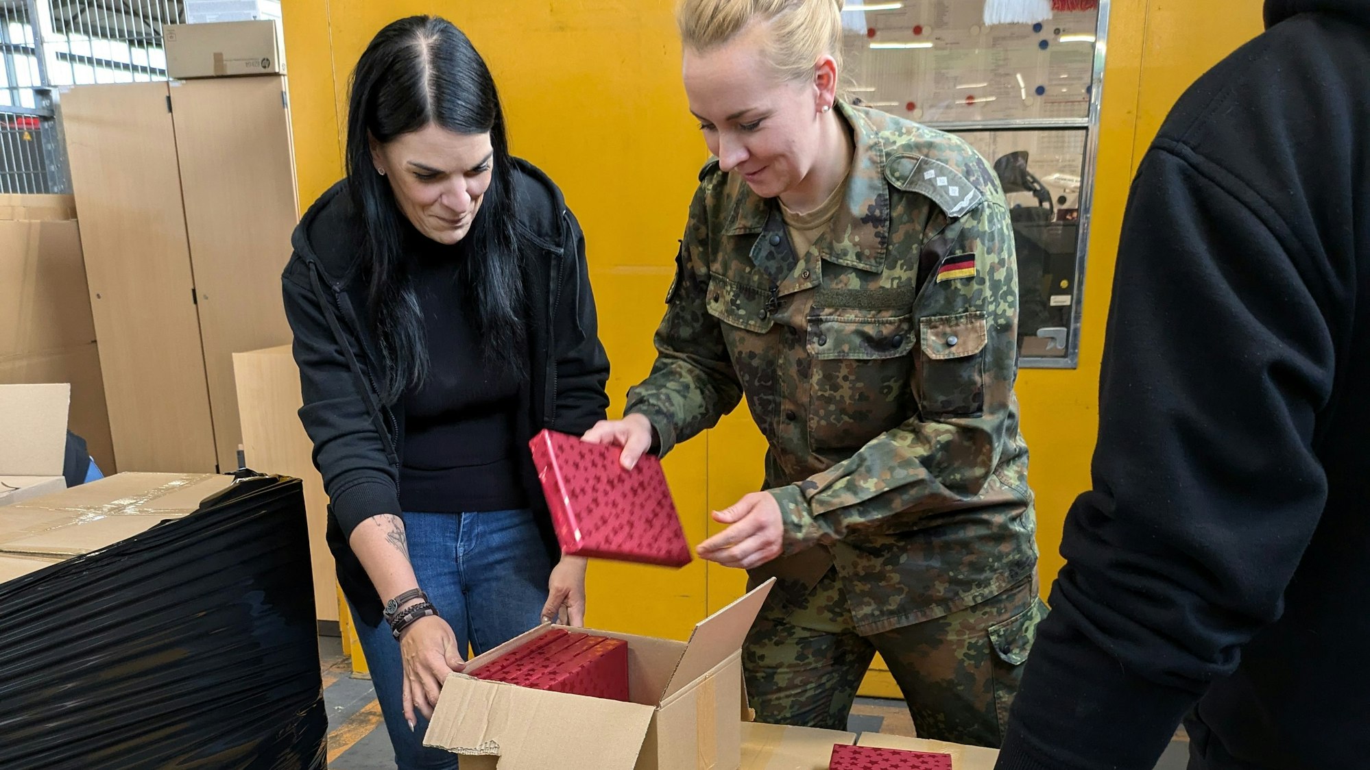 Hauptmann Nadine L. nimmt ein Geschenk, das in rotes Papier verpackt ist, und steckt es in ein Paket. Eine Zivil-Beschäftigte der Bundeswehr hält die Kiste auf.