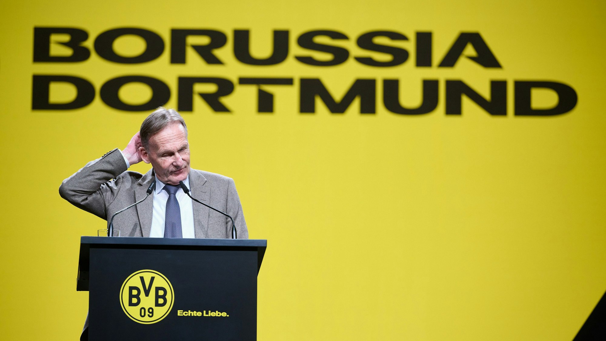 Hans-Joachim Watzke steht auf der BVB-Mitgliederversammlung am Rednerpult und kratzt sich am Kopf.