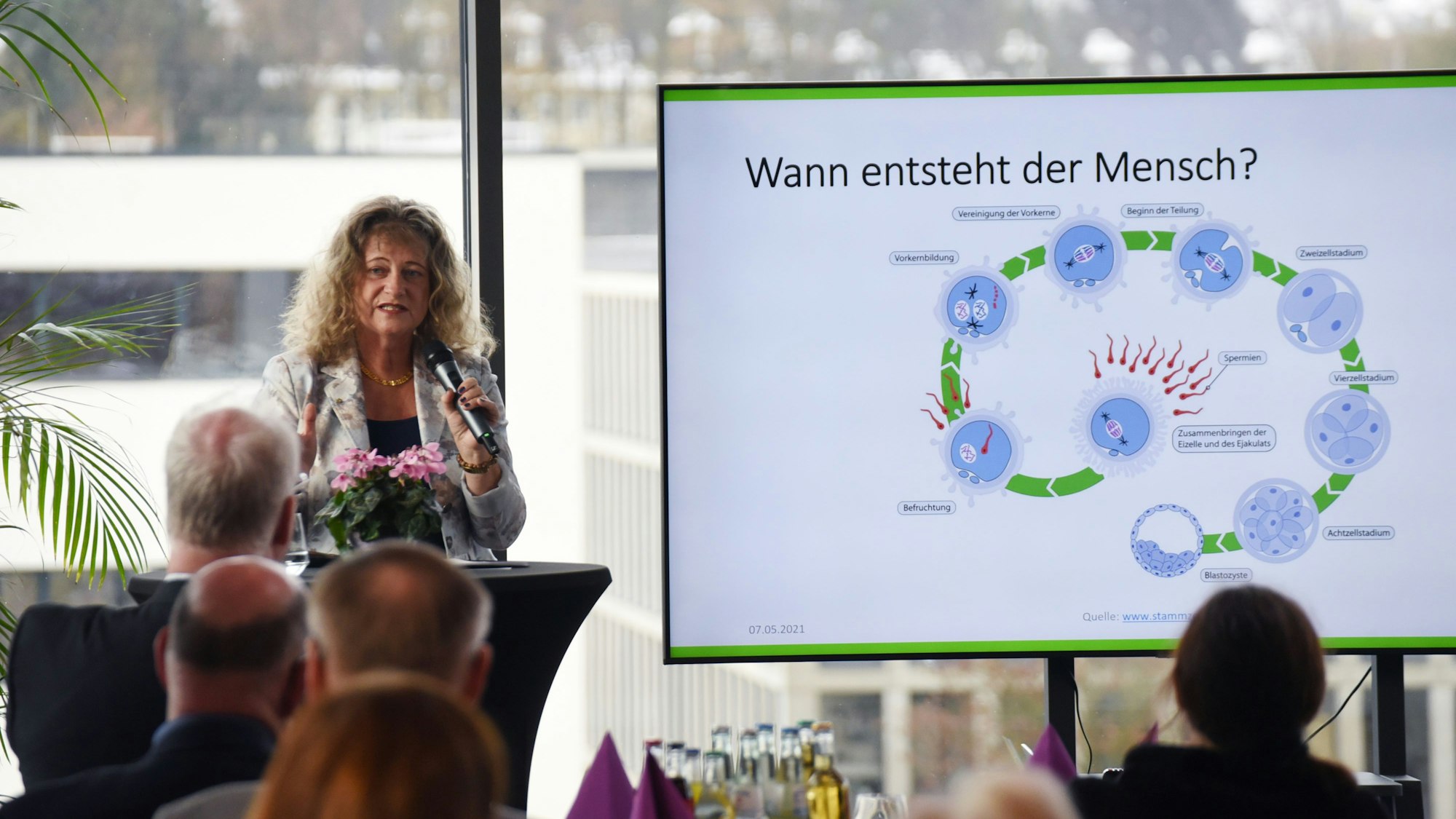 Eine Frau spricht in ein Mikrofon, daneben ein Bildschirm mit einer Grafik und der Frage: "Wann entsteht ein Mensch?"