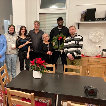 Die Band Polychrom posiert im Café International in Euskirchen hinter einem Tisch, auf dem ein Weihnachtsstern steht. Ein großer Mann mit Kappe hält einen Adventskranz, eine junge Frau formt mit der Hand die Rockgeste „Pommesgabel“.