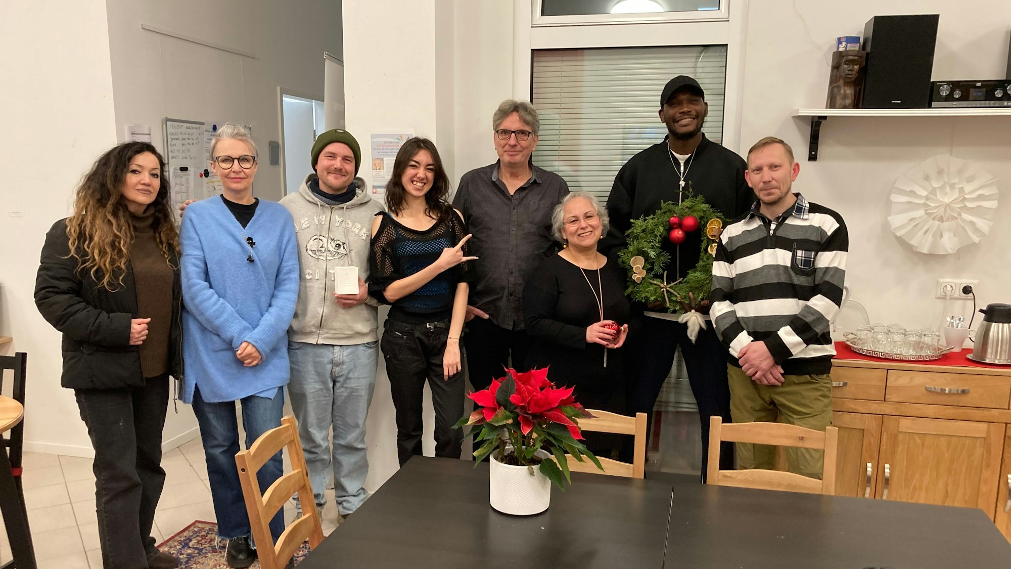 Die Band Polychrom posiert im Café International in Euskirchen hinter einem Tisch, auf dem ein Weihnachtsstern steht. Ein großer Mann mit Kappe hält einen Adventskranz, eine junge Frau formt mit der Hand die Rockgeste „Pommesgabel“.
