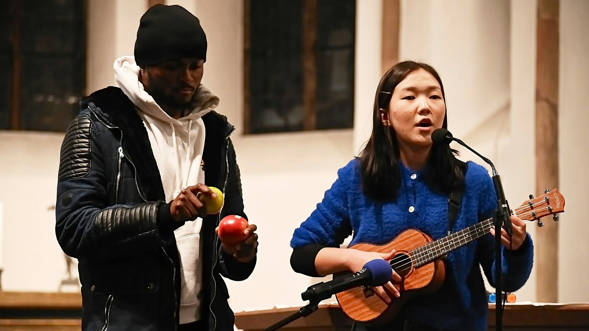 Die 14-jährige Enuun singt und spielt Ukulele, neben ihr steht ein dunkelhäutiger Mann, der zwei Rhythmusinstrumente in den Händen hält.