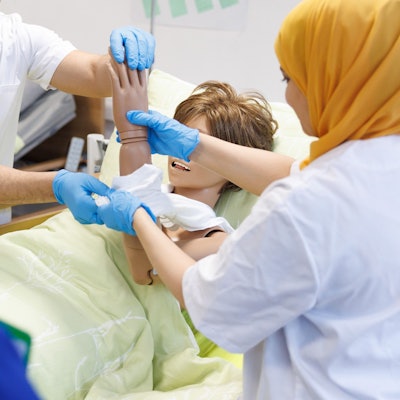 Eine Frau mit gelbem Kopftuch hält den Arm einer Körperpuppe und führt ihn durch ein T-Shirt. Ihr gegenüber steht ein Mann und assistiert ihr.