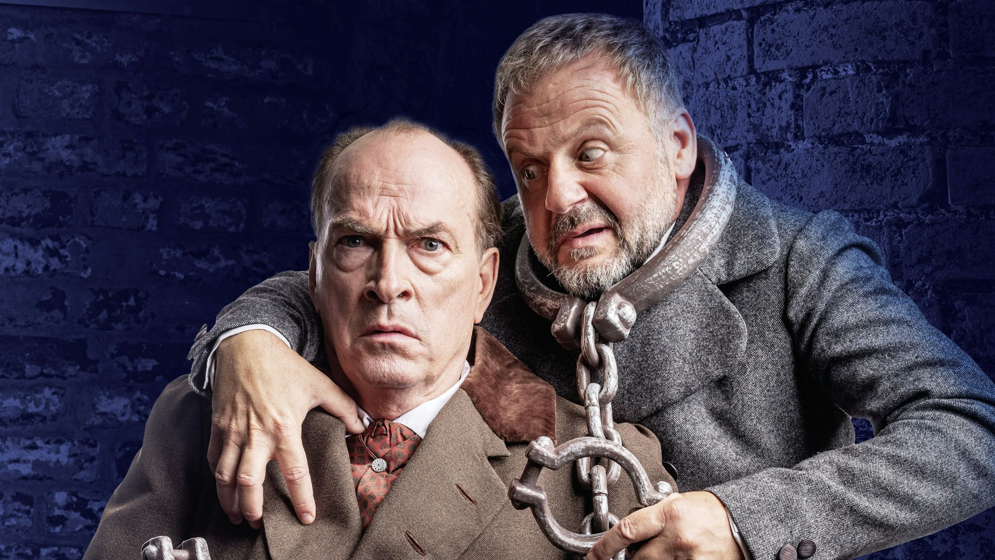 Knaup trägt einen braunen Mantel und schaut verdutzt, Samuel Finzi trägt eine schwere Eisenkette um den Hals und hat seinen Arm um Knaup gelegt.