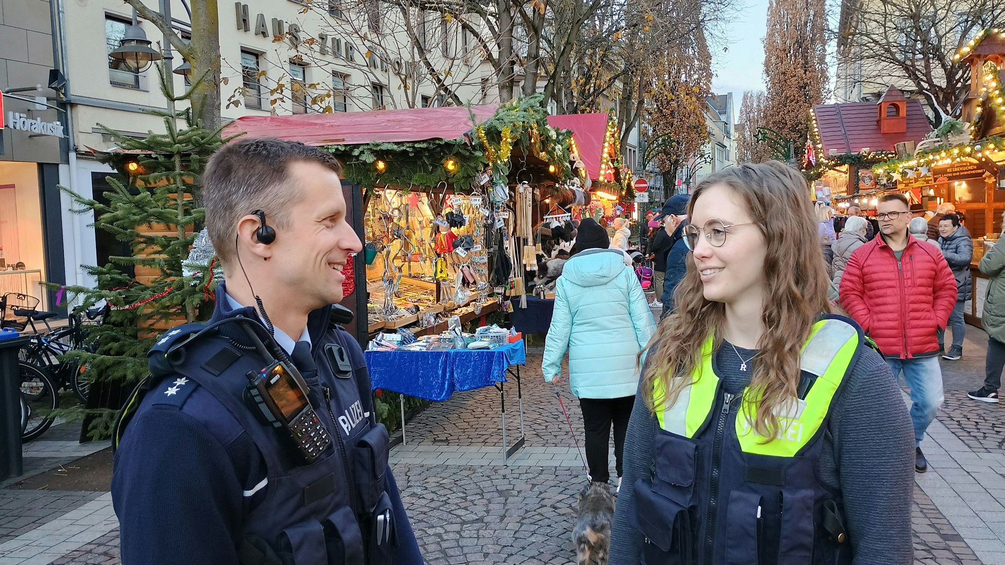 Die beiden tragen Polizeiwesten. Im Hintergrund die Stände des Weihnachtsmarktes und einige Passanten.