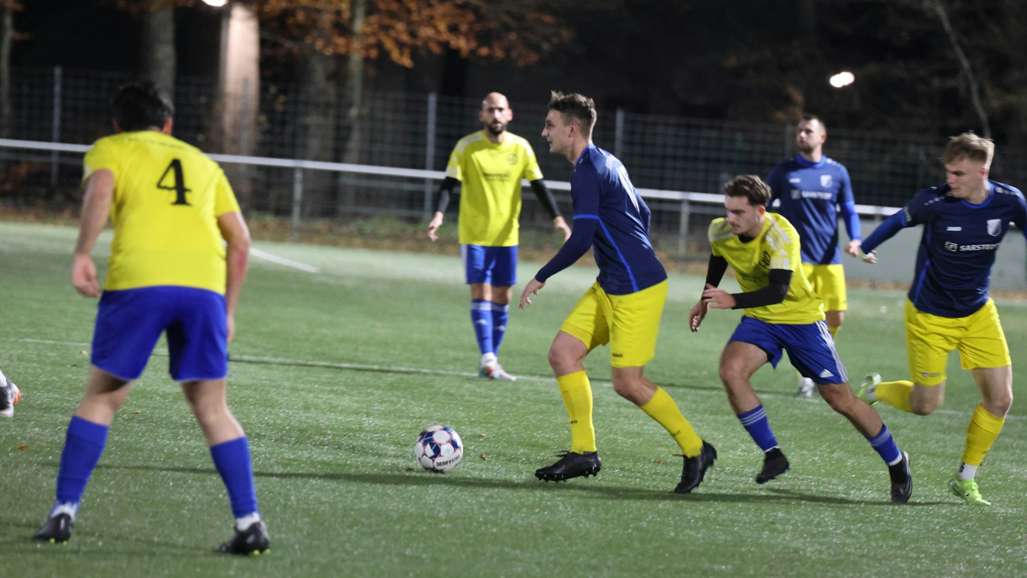 Zwei Fußballteams spielen auf einem Rasenplatz im Flutlicht abends gegeneinander.