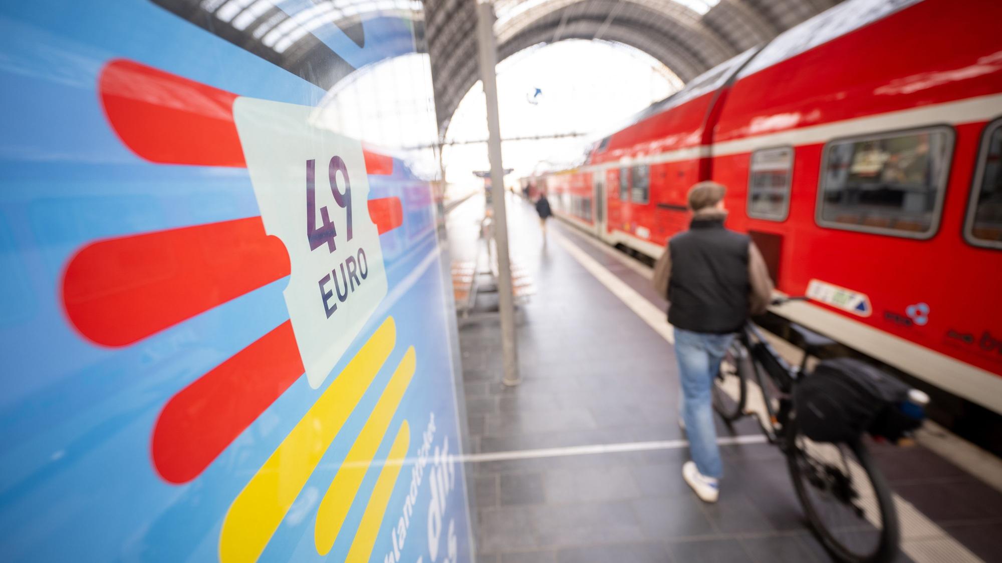 Ein Mann geht am Frankfurter Hauptbahnhof mit seinem Fahrrad zwischen der Werbung für das Deutschlandticket und einer Regionalbahn entlang.