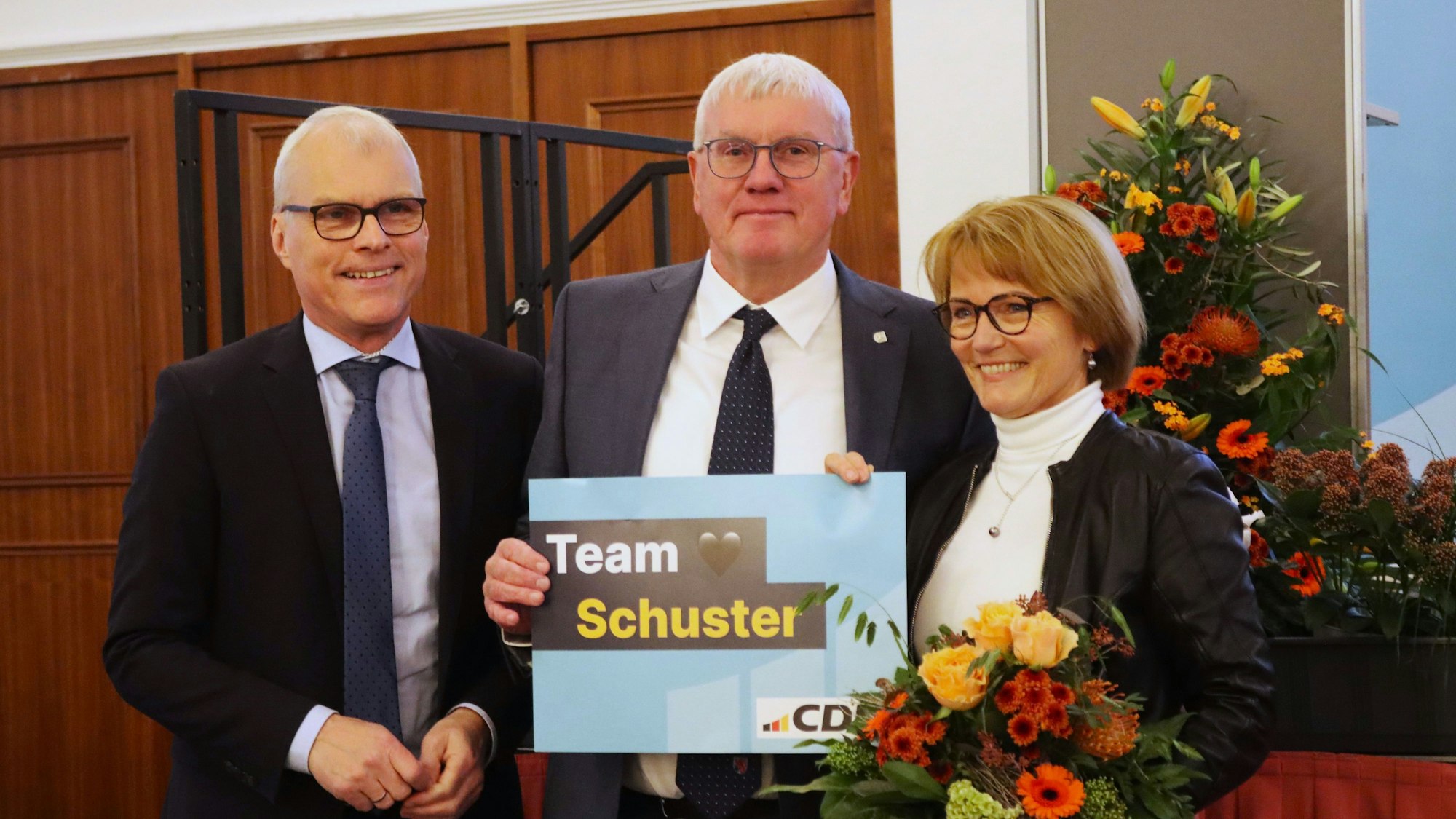 Zwei Männer und eine Frau; der Mann in der Mitte hält ein Schild mit der Aufschrift Team Schuster, die Frau einen Blumenstrauß.