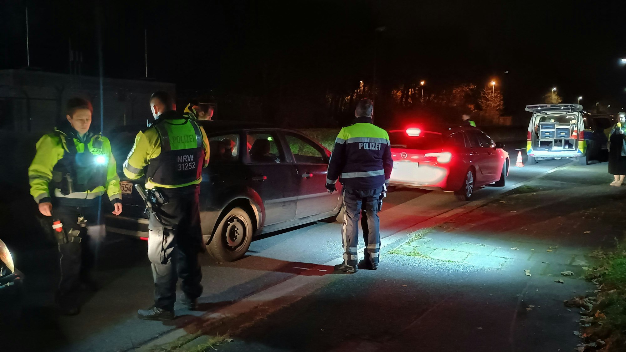 Auf dem Bild sind Polizisten zu sehen, die Autos kontrollieren. Es ist dunkel.