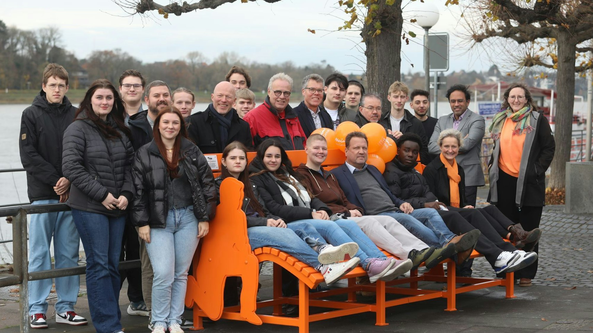 Viele Menschen stehen am Rhein hinter einer orangen Bank beziehungsweise sitzen auf ihr.