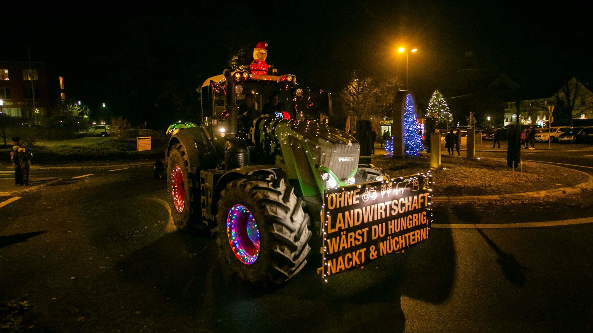 Ein beleuchteter Trecker, weihnachtlich geschmückt und mit einem Demo-Schild versehen, fährt auf einer Straße.