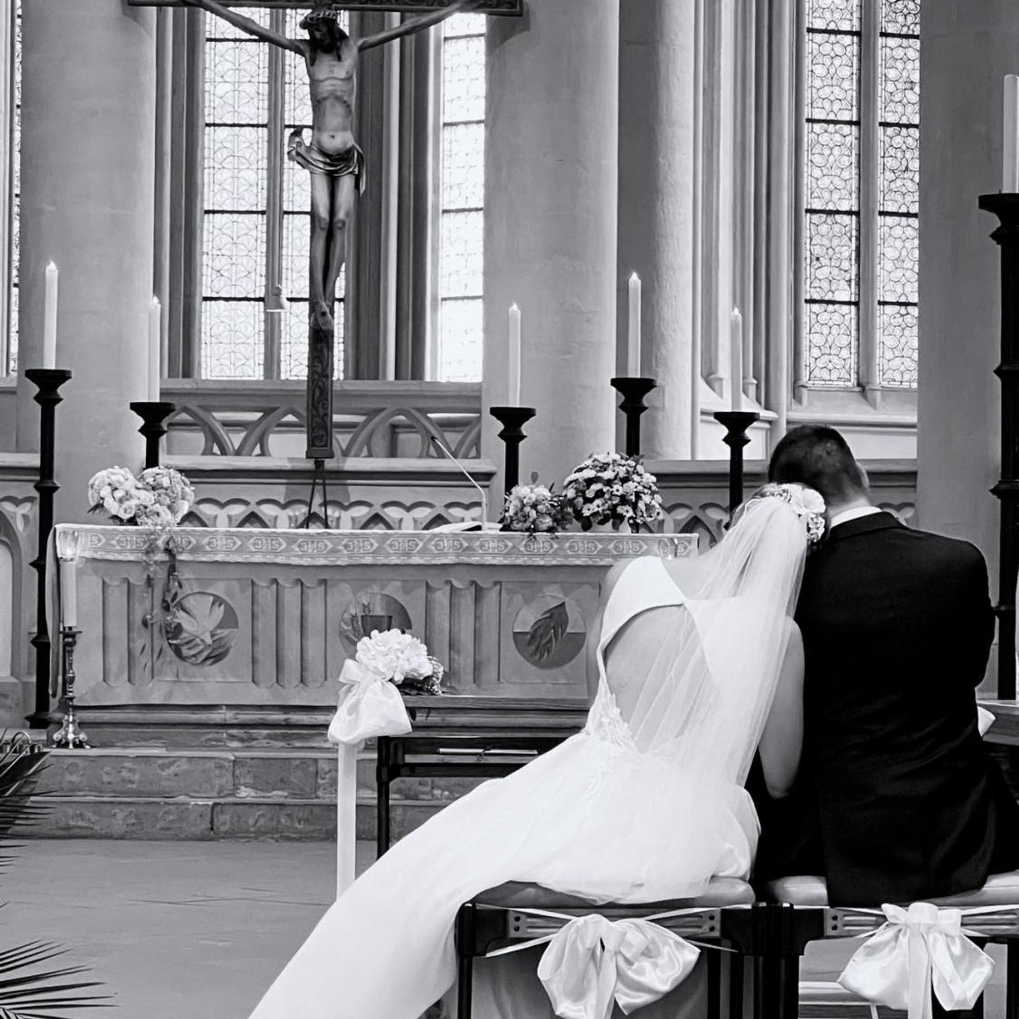 Braut und Bräutigam sitzen auf Hockern in einer Kirche. Die Braut legt ihren Kopf auf die Schulter des Bräutigams.