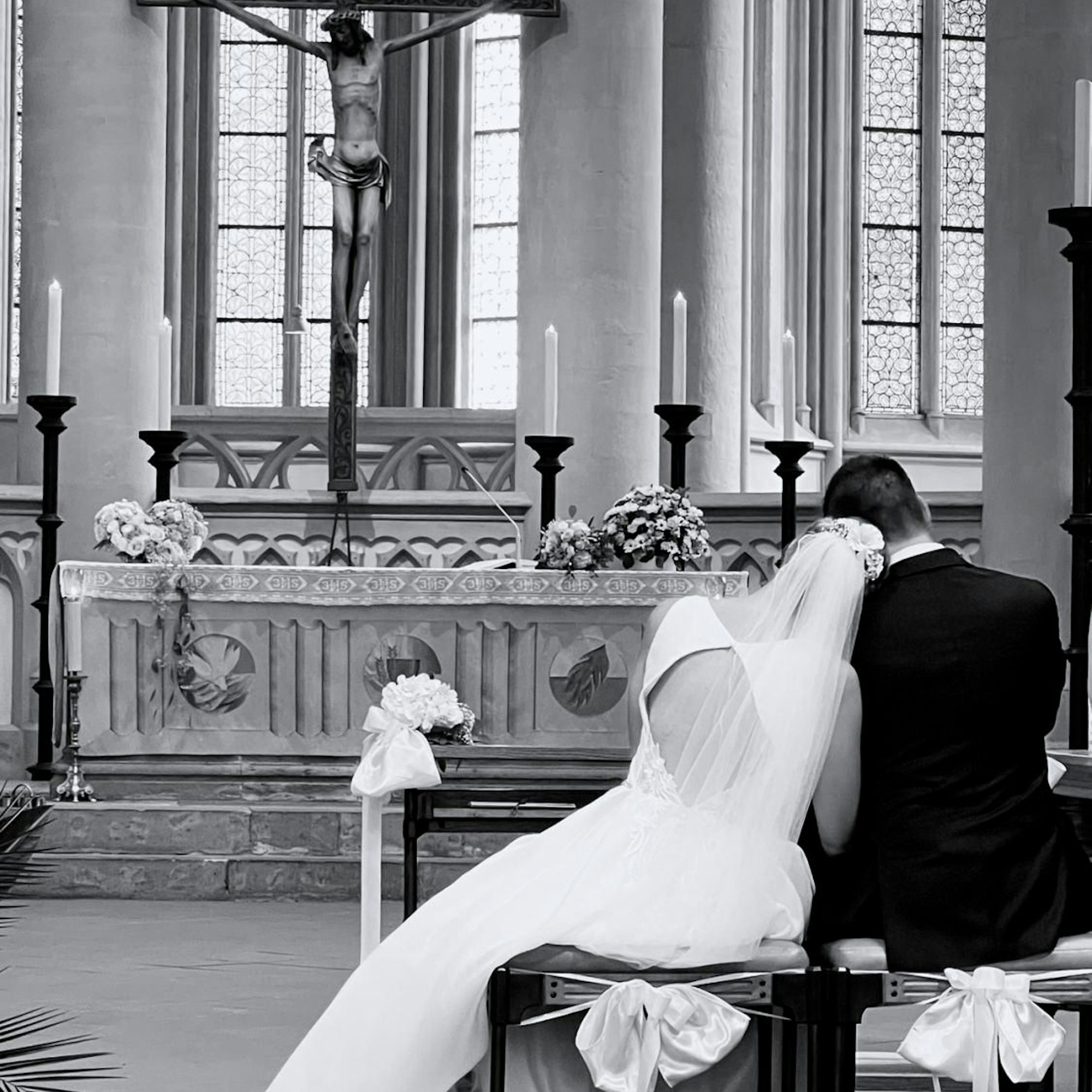 Braut und Bräutigam sitzen auf Hockern in einer Kirche. Die Braut legt ihren Kopf auf die Schulter des Bräutigams.