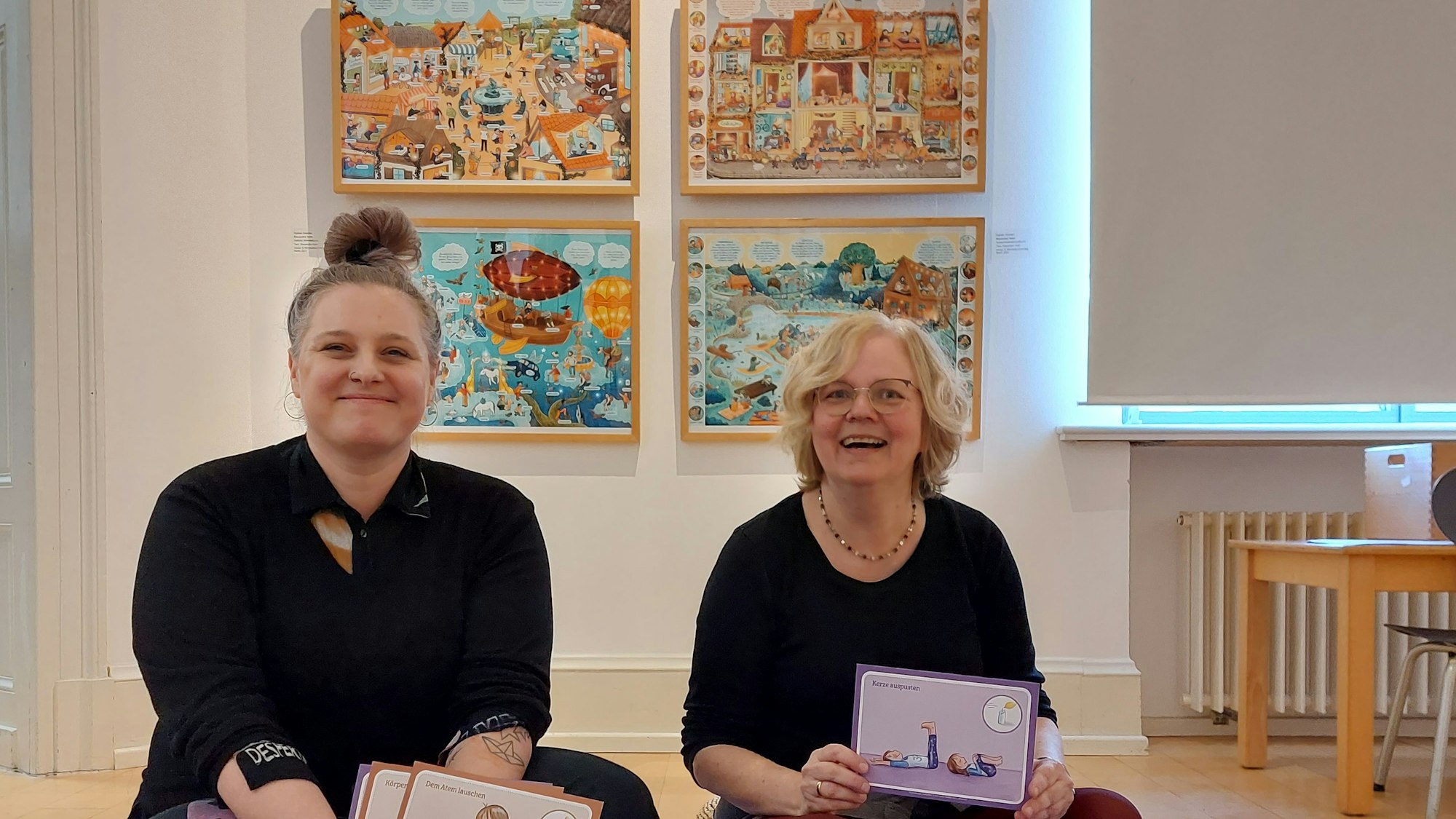 Zwei Frauen sitzen auf dem Boden, im Hintergrund hängen Bilderbuchillustrationen an der Wand.