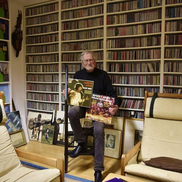 Ein älterer Herr mit Brille sitzt in einem Raum und hält zwei Plattencover in den Händen. Im Hintergrund sind haufenweise CDs in Regalen zu sehen, die bis an die Decke ragen.