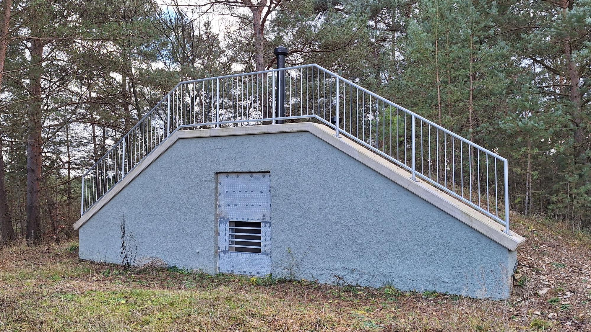 Ein trapezförmiges Gebäude, das in einem Wald steht, hat an der Forderseite eine Gittertür.