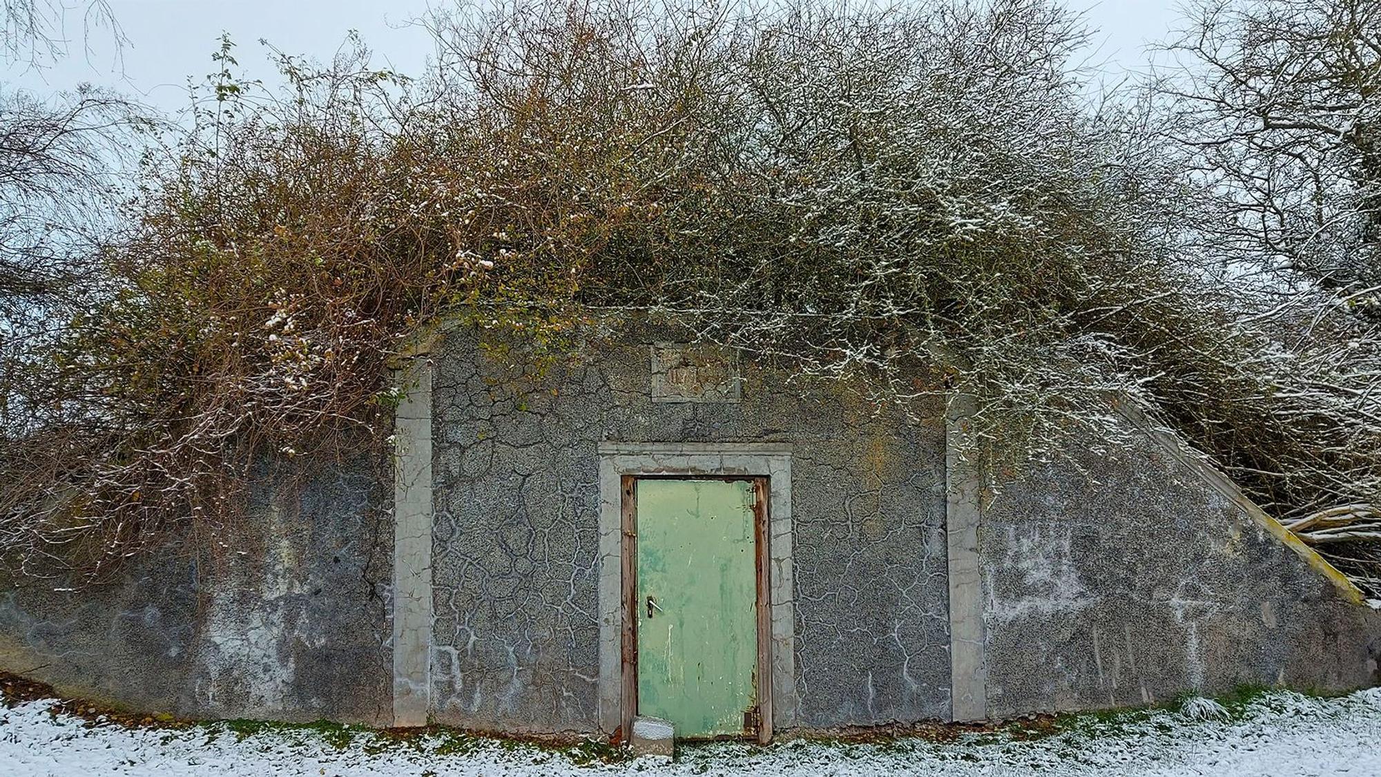 Außenansicht eines ehemaligen Wasserspeichers. Vor dem bunkerartigen Gebäude mit einer Tür liegt Schnee.