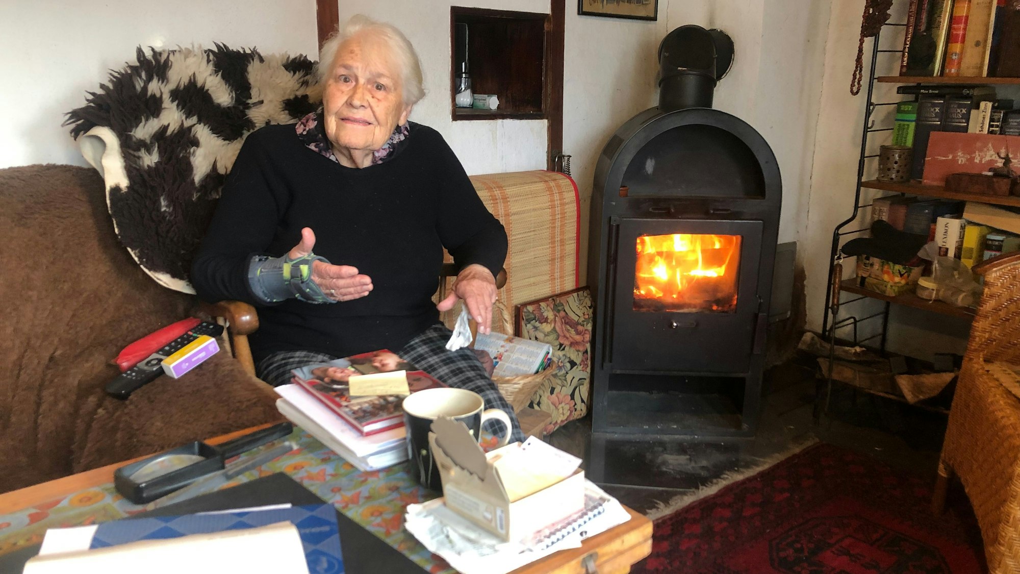 Eine Seniorin sitzt auf einer Couch, hinter ihr brennt Feuer in einem Kaminofen.