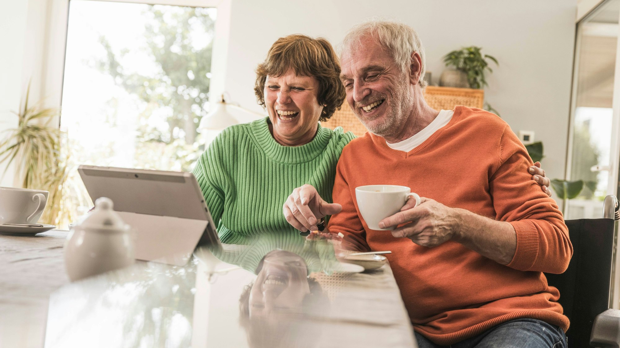 Eine ältere Frau und ein älterer Mann sitzen lachend an einem Tisch vor einem Tablet