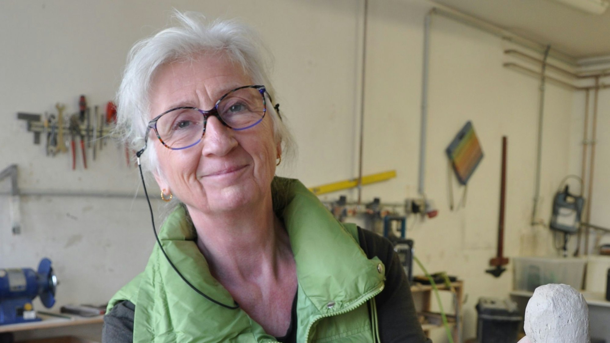 Die Steinmetzmeistern steht in ihrer Werkstatt und modelliert eine Figur. Sie trägt eine grüne Weste und eine Brille.