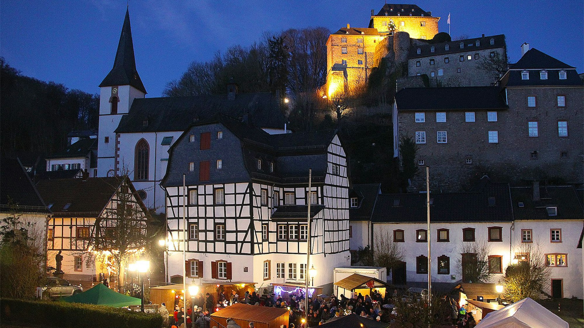Das Bild zeigt die erleuchtete Burg in Blankenheim. Im Vordergrund ist einer kleiner Weihnachtsmarkt zu sehen.