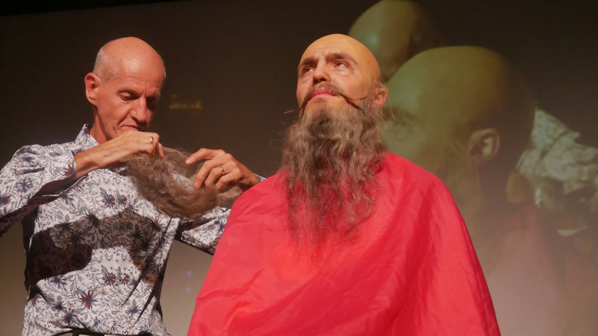 Ein Mann mit Glatze bekommt auf der Bühne einen Bart und eine Perücke angepasst.