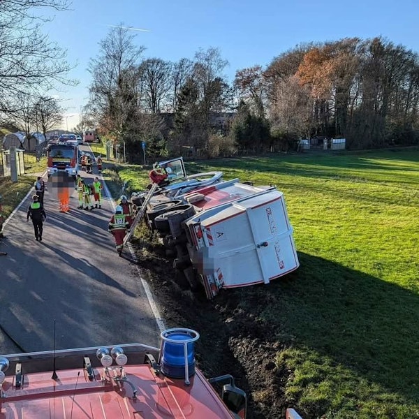 Ein Müllwagen liegt auf der Seite in einer Wiese, Feuerwehrleute steigen auf einer Leiter zur Fahrertür hinauf.