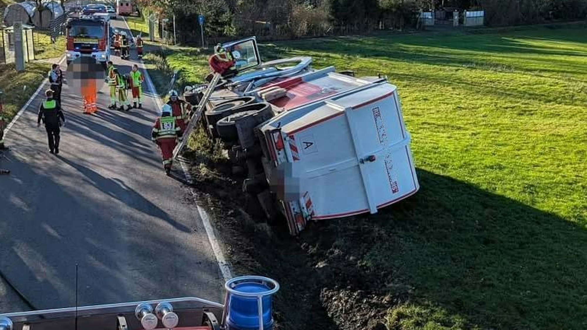Ein Müllwagen liegt auf der Seite in einer Wiese, Feuerwehrleute steigen auf einer Leiter zur Fahrertür hinauf.