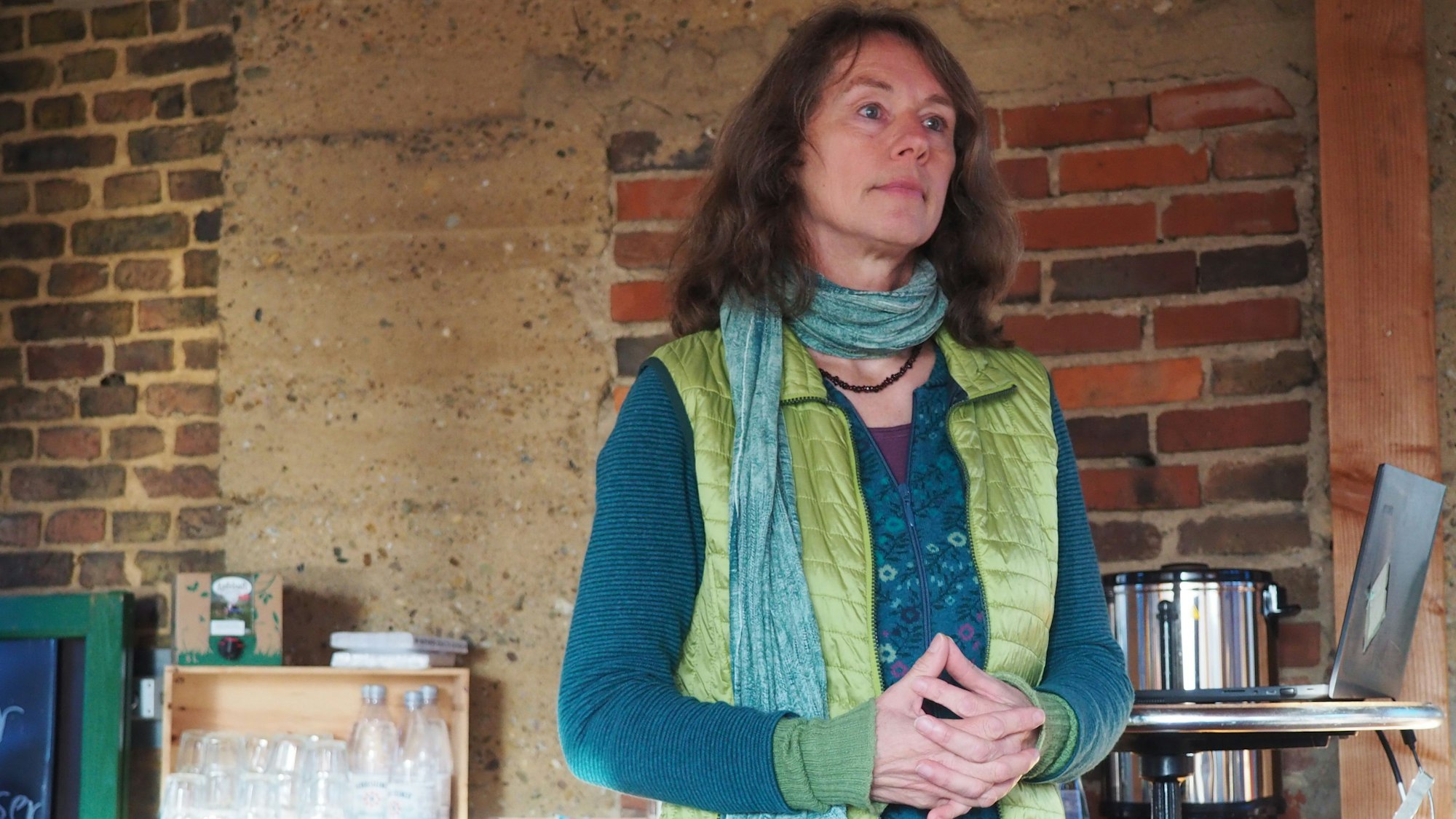 Elke Sprunkel steht vor einer Ziegelwand. Sie trägt ein türkises Oberteil unter einer grünen Weste.