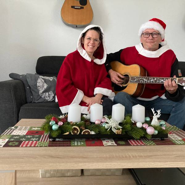 Rainer Behr und Susann Behr-Lauscher sitzen in weihnachtlichen Ponchos an einem Tisch, auf dem Adventskerzen stehen. Behr hält eine Gitarre.