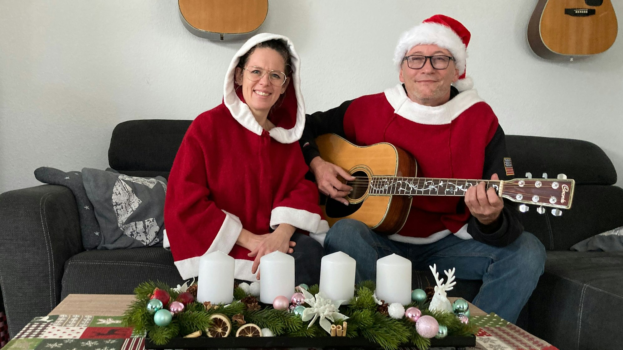 Rainer Behr und Susann Behr-Lauscher sitzen in weihnachtlichen Ponchos an einem Tisch, auf dem Adventskerzen stehen. Behr hält eine Gitarre.