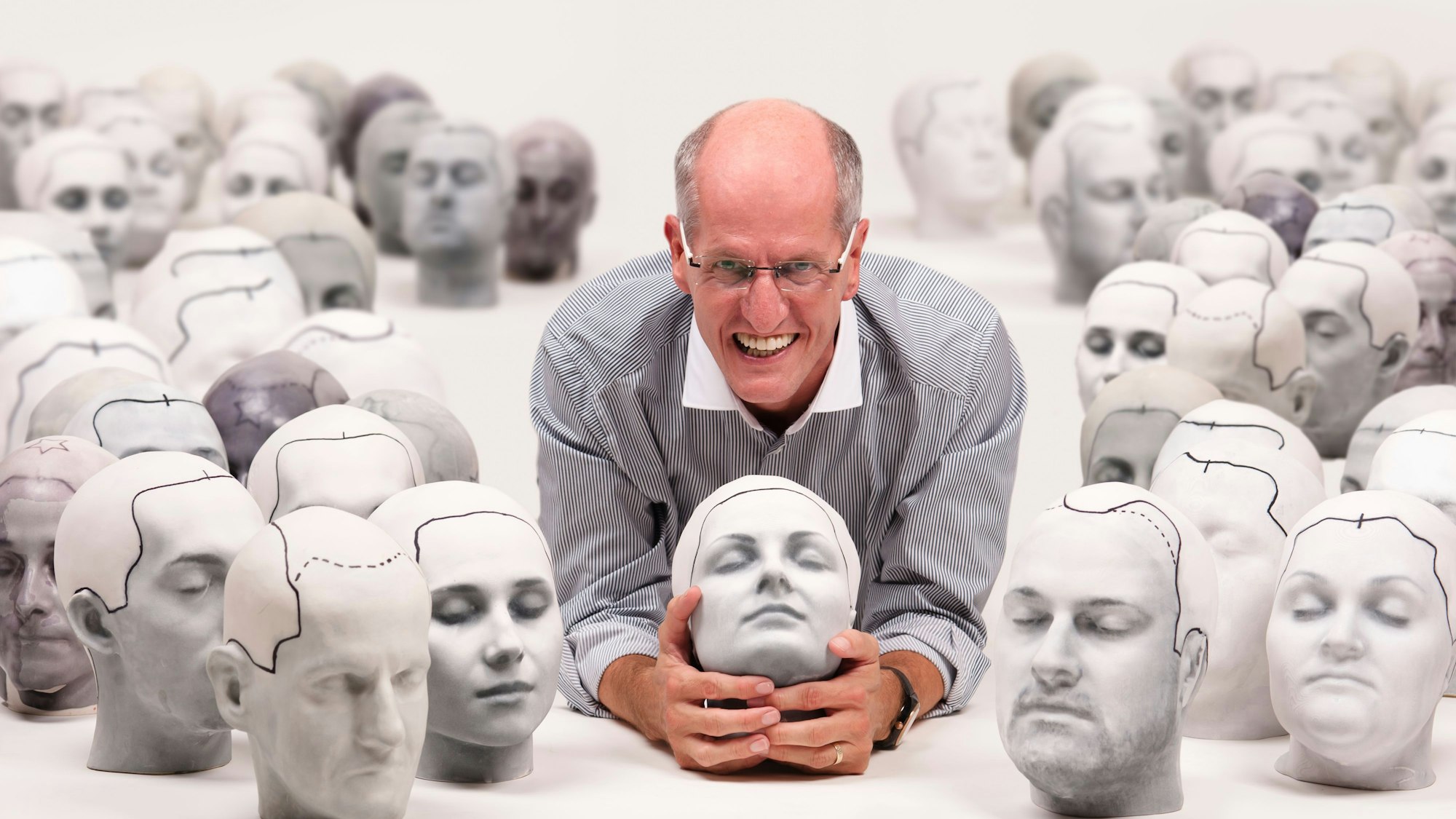 Ein Mann liegt am Boden. Er hat einen künstlichen Frauenkopf in der Hand und ist umgeben von vielen weiteren Kunstköpfen.