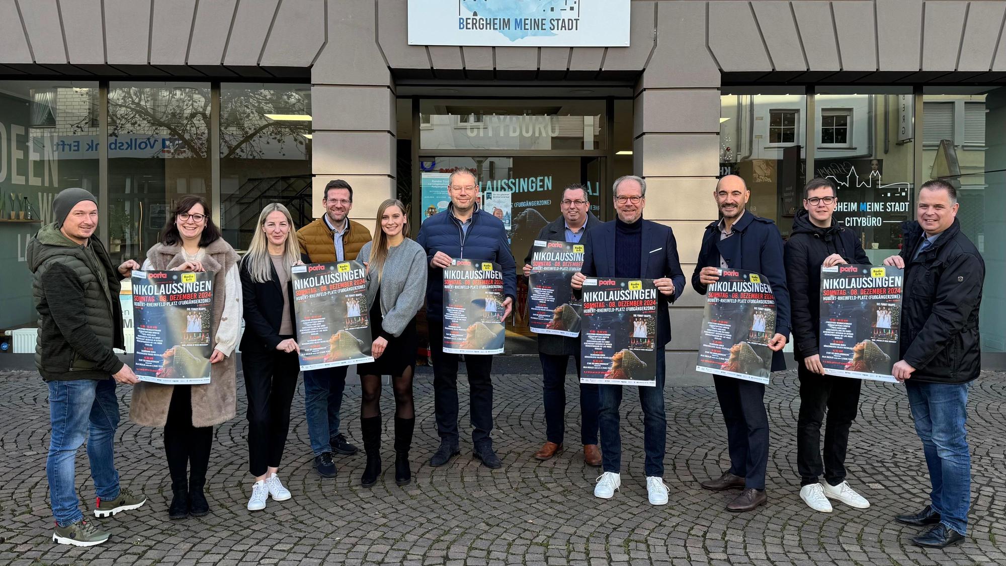 Zu sehen sind verschiedene Akteure aus Stadt und Kultur, die Plakate für das Nikolaussingen hochhalten.