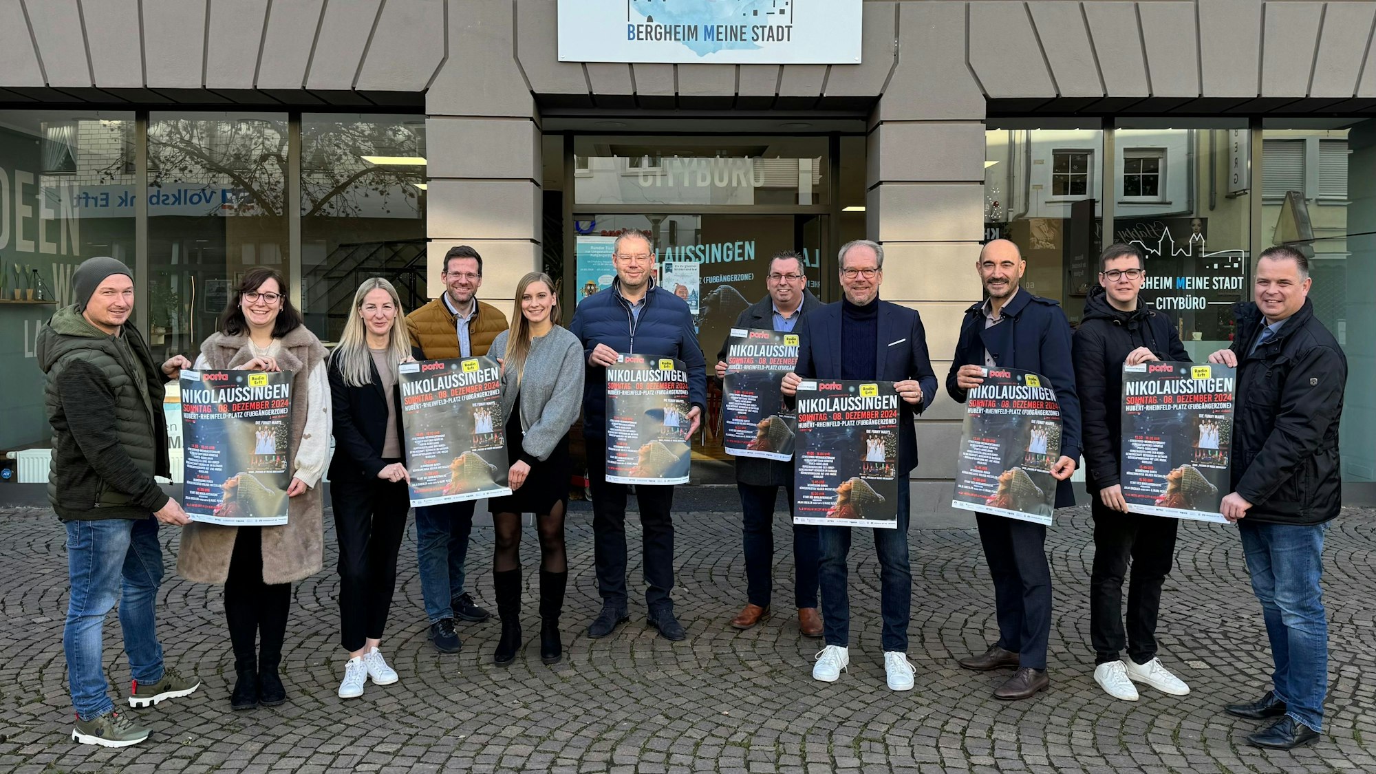 Zu sehen sind verschiedene Akteure aus Stadt und Kultur, die Plakate für das Nikolaussingen hochhalten.