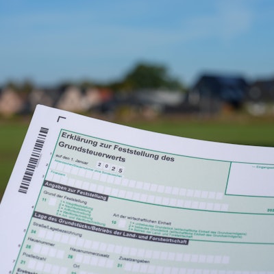 Ein Erklärung zur Feststellung des Grundsteuerwerts für das Jahr 2025 wird vor einer Wohnsiedlung hochgehalten.
