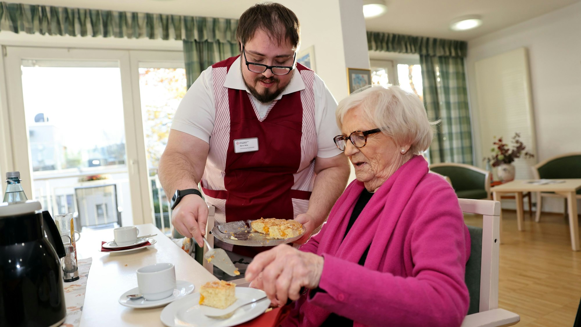 Den Kontakt mit Menschen schätzt Marco Vella bei seiner Arbeit sehr. Er serviert eine Dame in einer pinkfarbenen Strickjacke ein STück Kuchen.