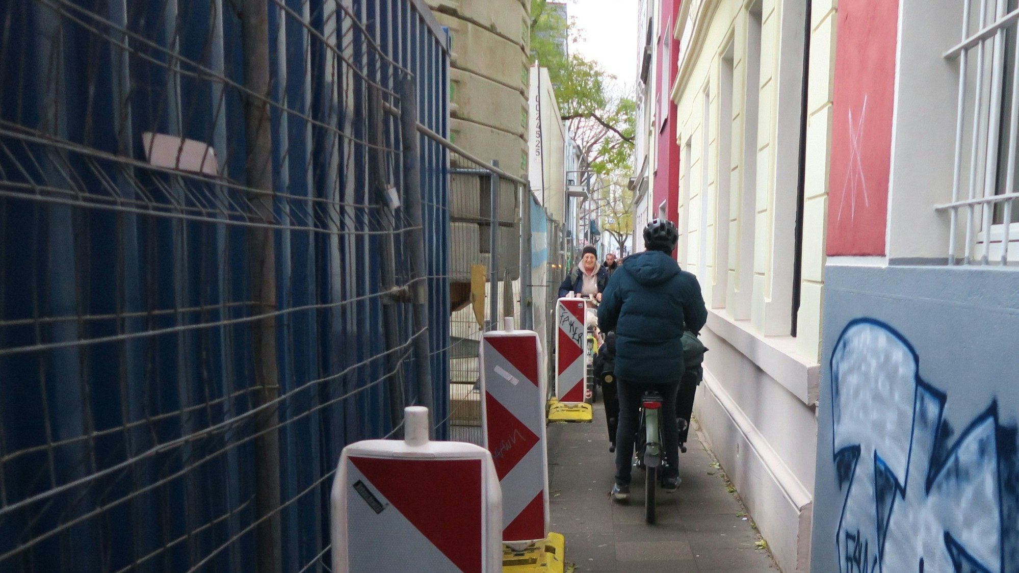 Auf einem sehr schmalen Gehweg fährt ein Mann mit Lastenrad, während ihm Fußgänger entgegenkommen.