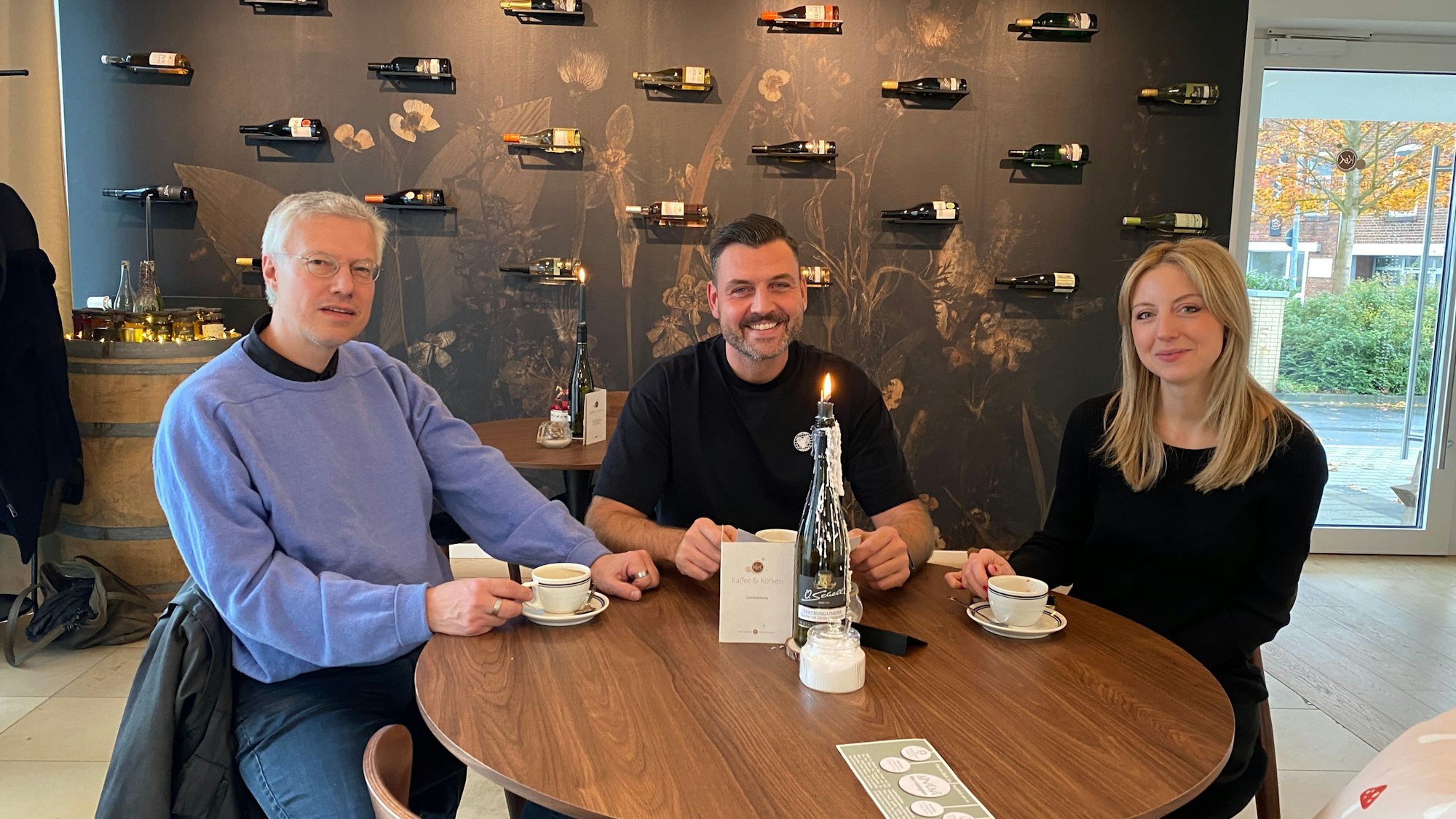 Janine Munder, Andŕe Schiebahn und Oliver Schell sitzen an einem Tisch, vor ihnen jeweils eine Tasse Kaffee. Im Hintergrund sind Weinflaschen auf Regalteilen an einer Wand zu sehen.