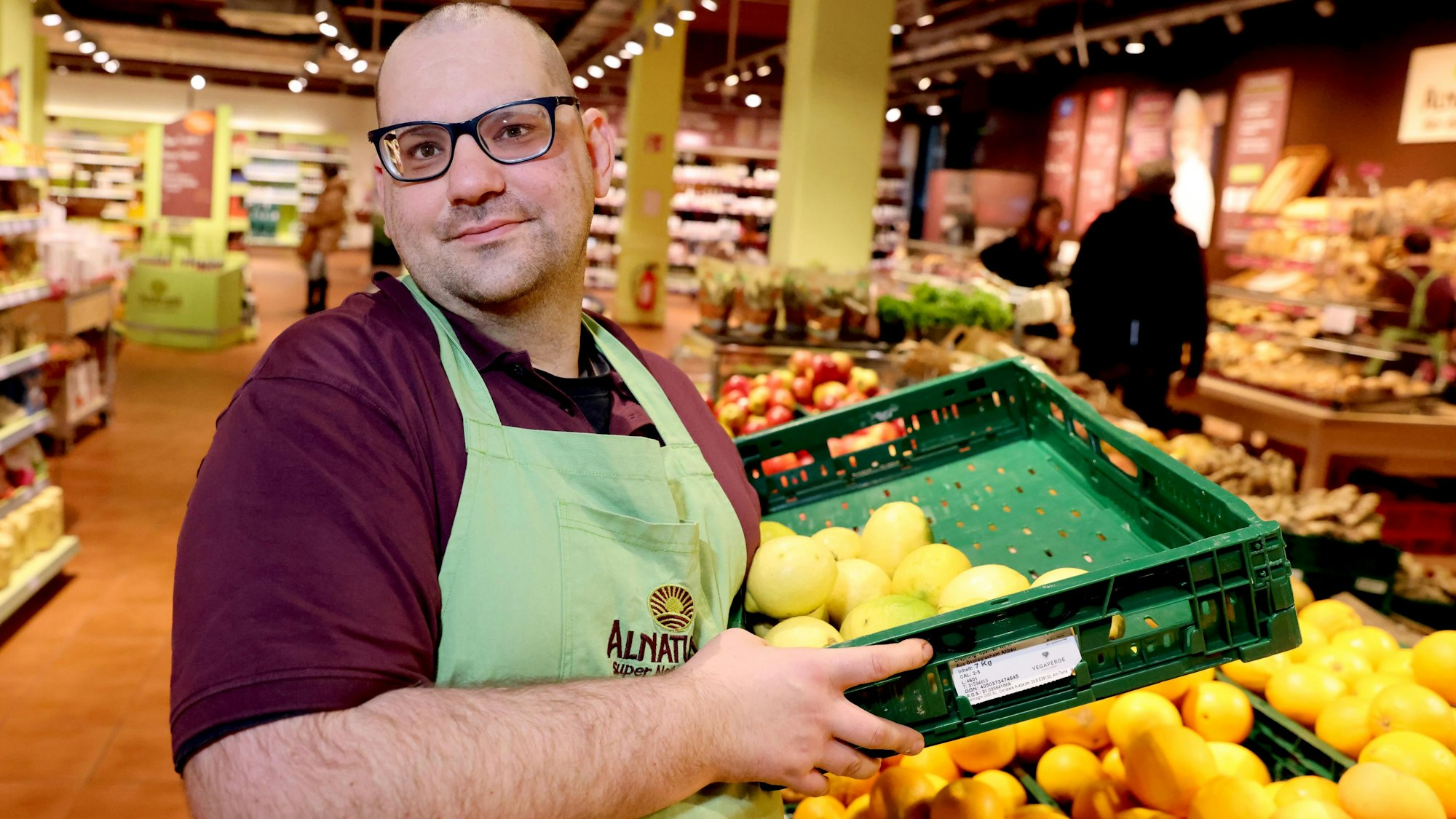 Die Verantwortung für das Auffüllen des Obst- und Gemüsestandes ist ein Aufgabenbereich von Paul Adamiuk. Er steht mit einer halbvollen Zitronenkiste am Obsstand.