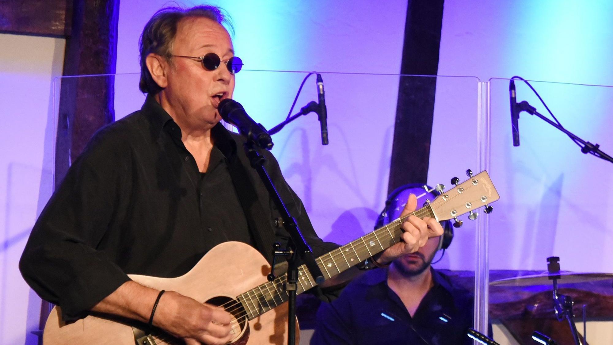 Ein Mann mit Sonnenbrille und Gitarre singt in ein Mikrofon.