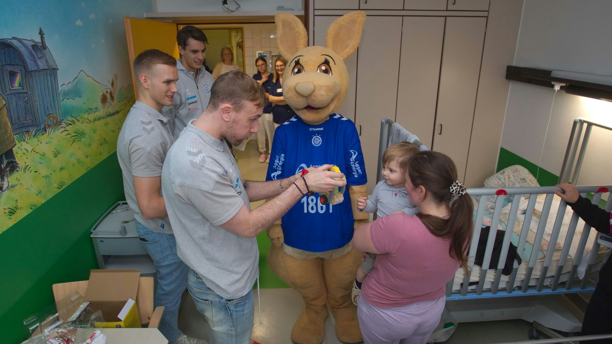 Drei Handballer überreichen in einem Krankenzimmer einem kleinen Jungen ein Spielzeug.