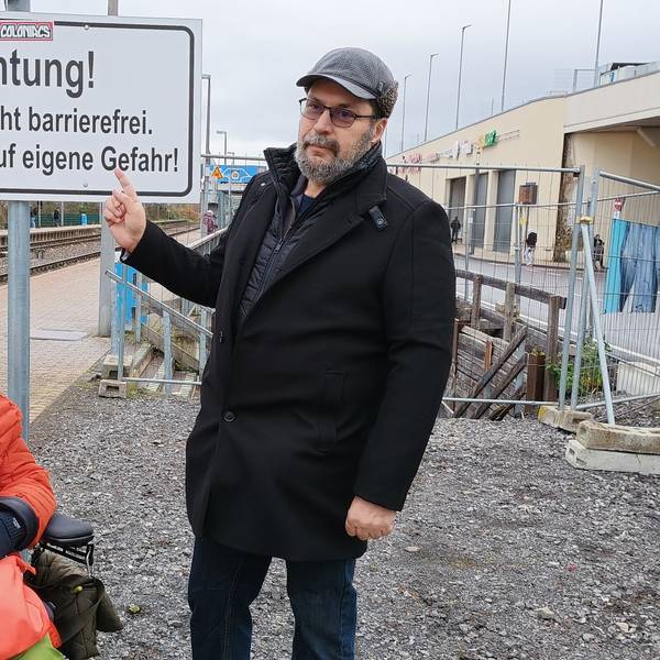 Dulgerakis zeigt mit dem Finger auf ein Schild mit der Aufschrift „Achtung! Rampe nicht barrierefrei. Benutzung auf eigene Gefahr!“