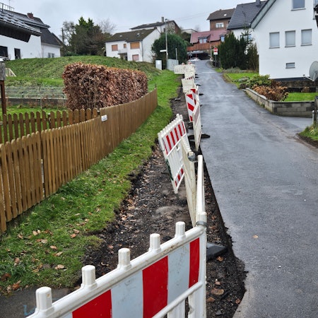 Das Bild zeigt eine Anwohnerstraße mit einer Baustellenabsperrung auf der linken Seite. Dort fehlt die letzte Asphaltschicht.