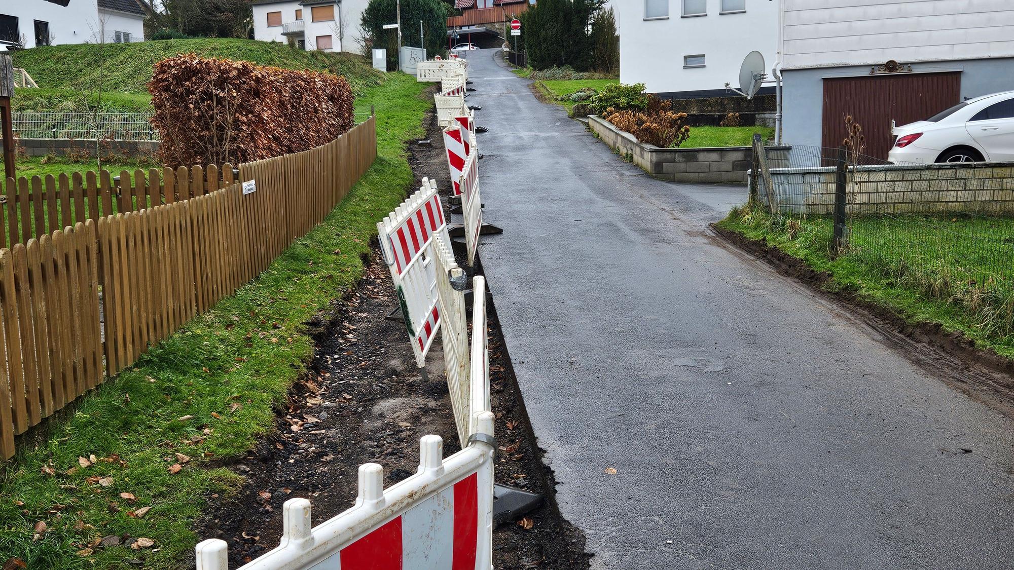 Das Bild zeigt eine Anwohnerstraße mit einer Baustellenabsperrung auf der linken Seite. Dort fehlt die letzte Asphaltschicht.