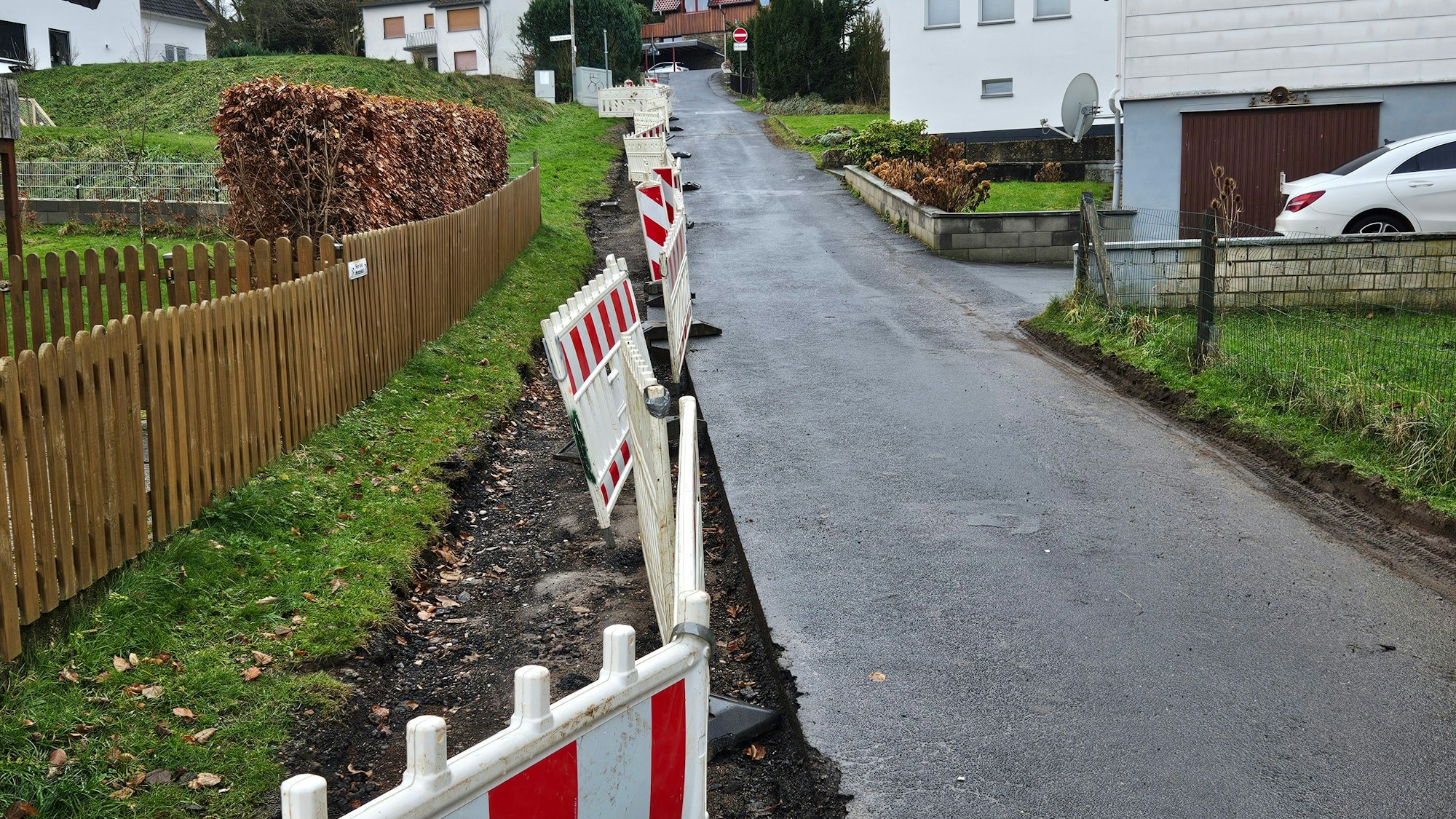 Das Bild zeigt eine Anwohnerstraße mit einer Baustellenabsperrung auf der linken Seite. Dort fehlt die letzte Asphaltschicht.
