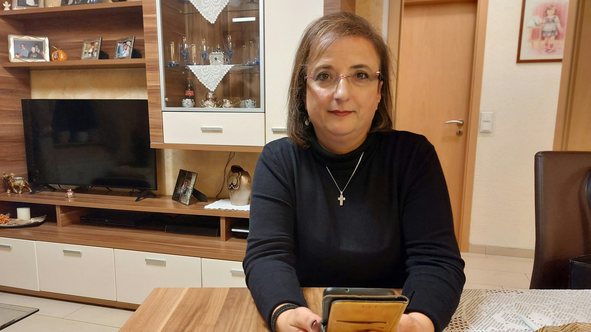 Eine Frau sitzt an einem Tisch in ihrer Wohnung. Sie hält ihr Mobiltelefon in der Hand.