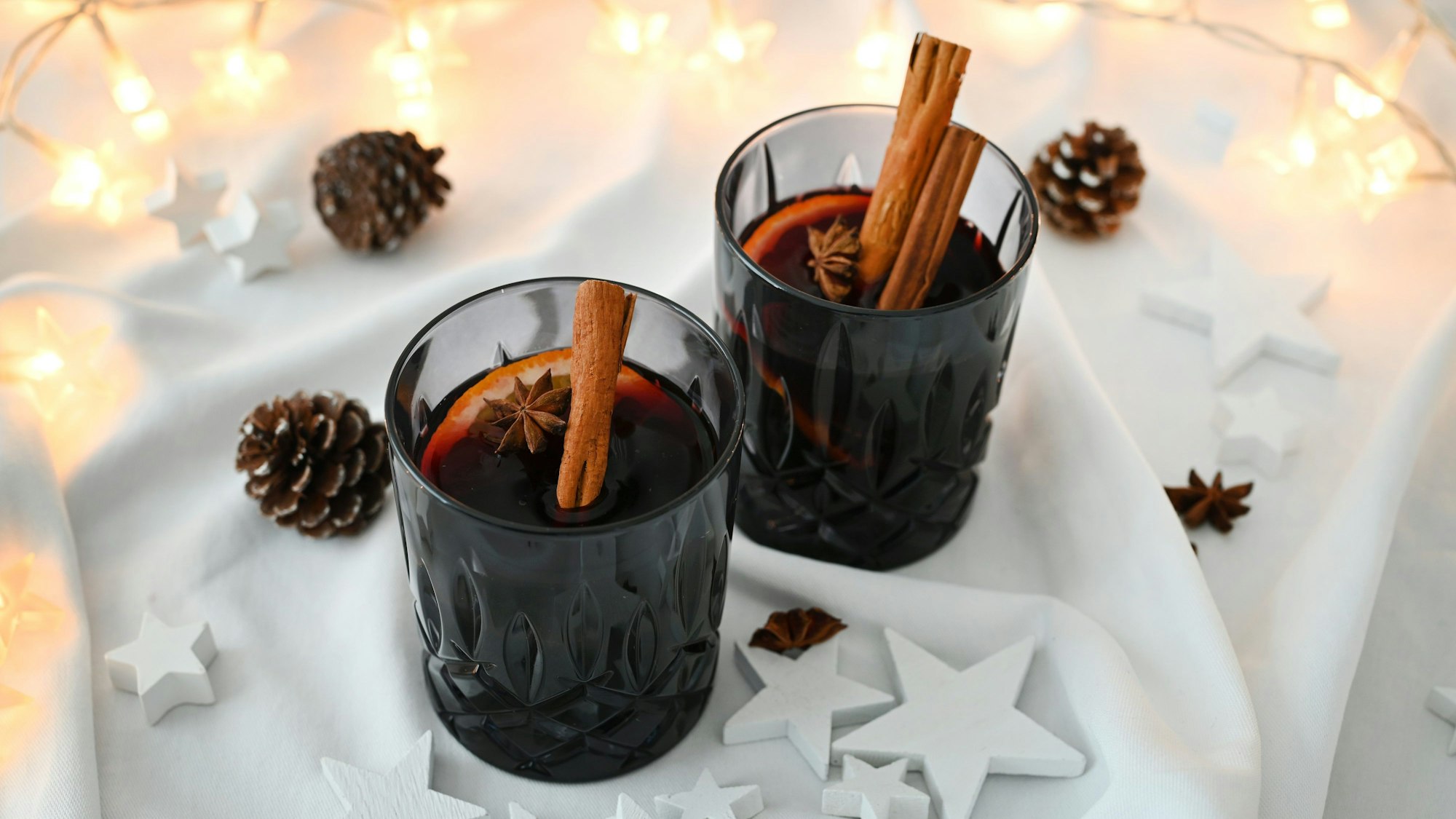 Zwei Gläser mit Glühwein stehen auf einer weißen Tischdecke. Aus den Gläsern steckt eine Stange Zimt und im Wein schwimmt ein Sternanis.