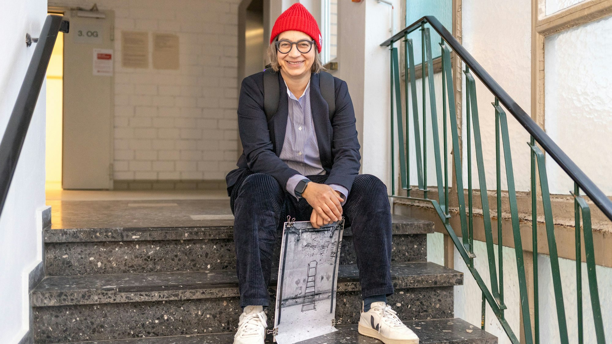 Claudia Mewaldt sitzt mit einem ihrer Bilder auf einer Treppe im Treppenhaus.