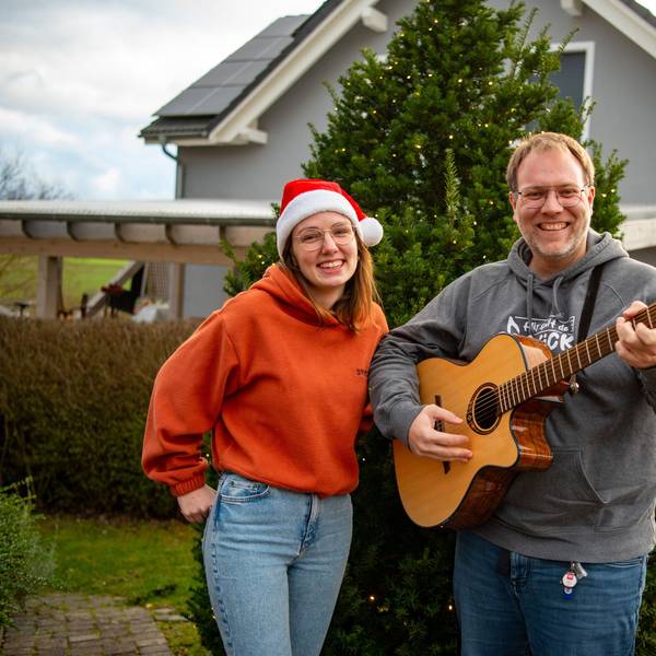 Michael Remer und Anna Jardin von der Sacro-Pop-Band Spirit stehen in Niederbettingen vor einem beleuchteten Tannenbaum. Anna Jardin trägt eine Weihnachtsmütze, einen orangen Pullover und helle Jeans. Michael Renner trägt grauen Pullover und dunkle Jeans und spielt Gitarre.