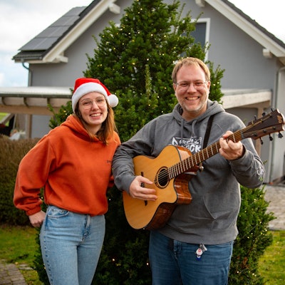 Michael Remer und Anna Jardin  von der Sacro-Pop-Band Spirit stehen in Niederbettingen vor einem beleuchteten Tannenbaum. Anna Jardin trägt eine Weihnachtsmütze, einen orangen Pullover und helle Jeans. Michael Renner trägt grauen Pullover und dunkle Jeans und spielt Gitarre.